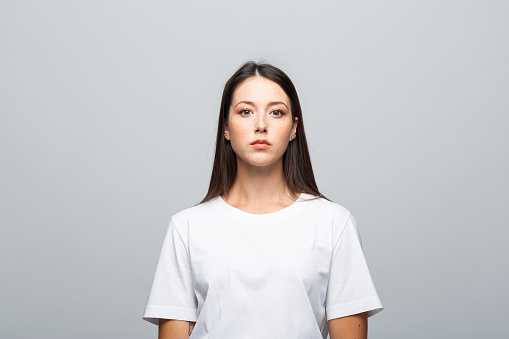 Portrait of beautiful young woman wearing white t-shirt, looking at camera. Studio shot, grey background.