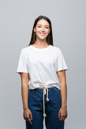 Portrait of beautiful young woman wearing white t-shirt and jeans, smiling at camera. Studio shot, grey background.