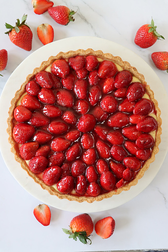 Stock photo showing uncut French strawberry tarte / torte with crispy, fluted pastry crust served on white plate. In the picture the golden, crisp pastry can be seen to be filled with strawberry halves that have been glazed with a strawberry jam.