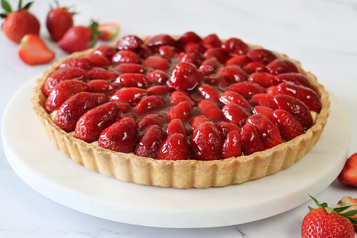 Stock photo showing uncut French strawberry tarte / torte with crispy, fluted pastry crust served on white plate. In the picture the golden, crisp pastry can be seen to be filled with strawberry halves that have been glazed with a strawberry jam.