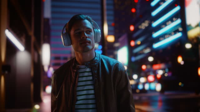 Portrait of Handsome Man Wearing Headphones Walking Through Night City Street Full of Neon Light. Smiling Stylish Man Listening to Music, Podcast, Talk Show. Dolly Tracking Medium Shot