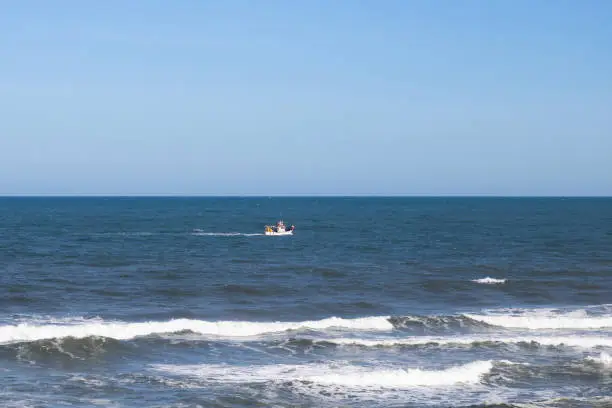 A small fishing boat sailing far away in the open Atlantic Ocean seen from the shore of Quiaios Beach, Portugal. Distant trawler and the horizon with clear blue sky