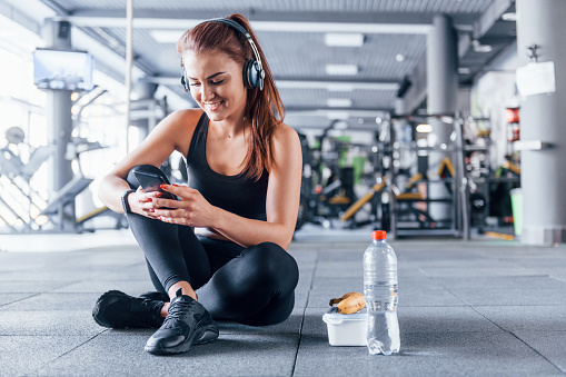 Young girl in sportive clothes and in headphones sitting in the gym at daytime with food.