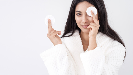 Skincare concept. Portrait of young happy asian woman in white bathrobe holding cotton pad covering her eye with it while standing on white background, smiling at camera and enjoying beauty routine