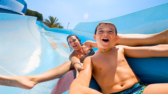 Happy cheerful caucasian son and mom laughing and shouting while sliding down fast on a water slide at aqua park. Family having fun on a sunny day in summer time. Lifestyle, vacation activities.