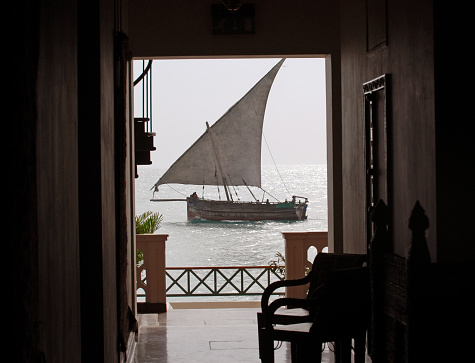 A passing Dhow (Arabic sailing ship) framed by the dark shadows of the entrance lobby in a  waterfront hotel, Stone Town, Zanzibar