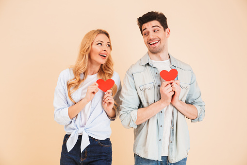 Portrait of lovely couple man and woman in basic clothing looking at each other with smile and holding two red paper hearts isolated over beige background