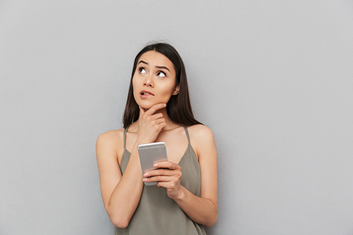 Portrait of a pensive asian woman holding mobile phone and looking away isolated over gray background