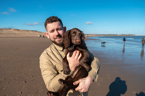 A portrait shot of a caucasian man, wearing casual clothing on a walk at the beach with his Brown Cocker Spaniel puppy on a sunny winters morning. He is looking and smiling at the camera while he carries his puppy.