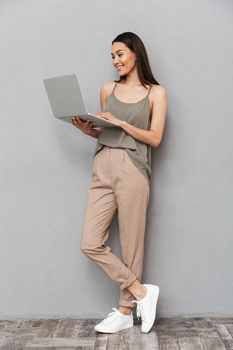 Full length portrait of a smiling asian woman using laptop computer isolated over gray background