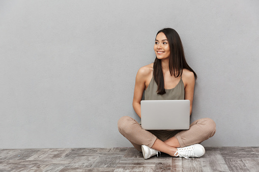 Portrait of a smiling asian woman sitting on a floor with laptop computer and looking away over gray background