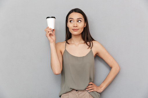 Portrait of a pretty asian woman holding take away coffee cup and looking away isolated over gray background
