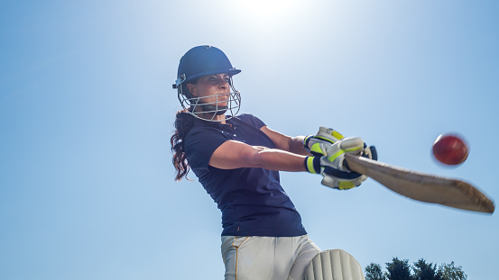 Low angle view of female batsman wearing protective gear and hitting the ball with a bat during the sunny day.