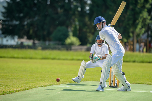 Male batsman hitting the ball while wicket-keeper standing behind stumps on the pitch.
