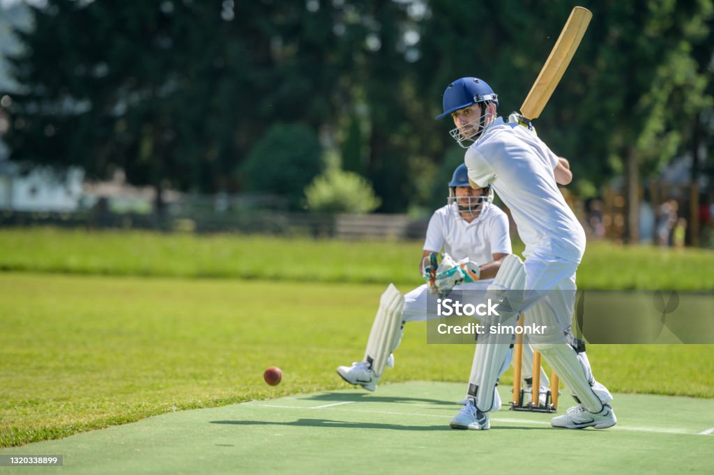 Batsman hitting ball on pitch Male batsman hitting the ball while wicket-keeper standing behind stumps on the pitch. Sport of Cricket Stock Photo