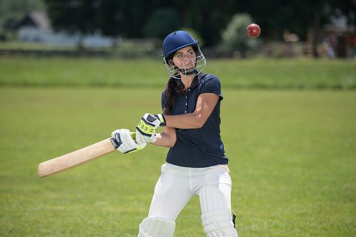 Female cricket player wearing protective gear and hitting the ball with a bat on the field.