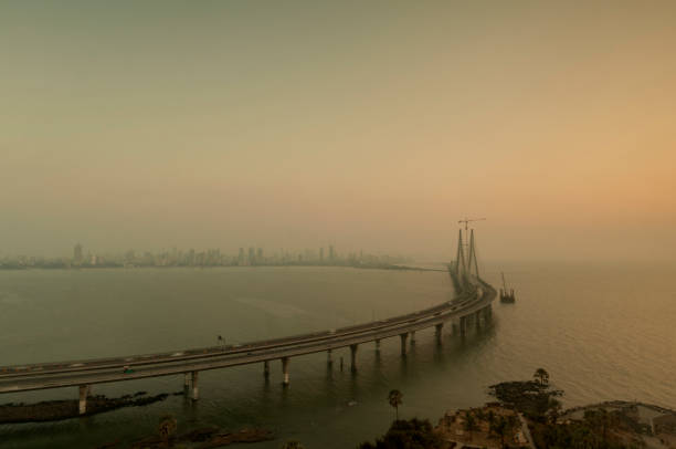 Sun setting over a smoggy Bandra Worli Sea Link Bridge, with the city skyline on the horizon.