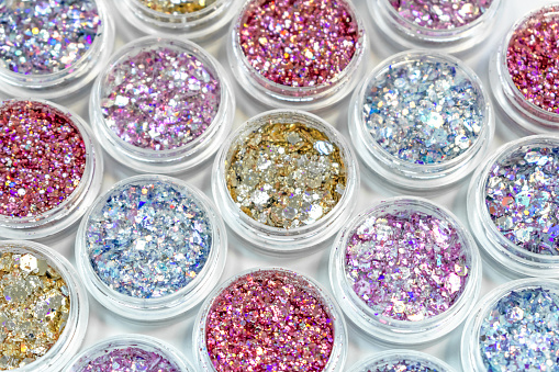 Blue, pink, gold and red glitter in jars on white background. Shiny holographic elements, sparkle. Beautiful shimmer for manicure, makeup, nail extension, design. Cosmetic products. Trendy delicate colors.