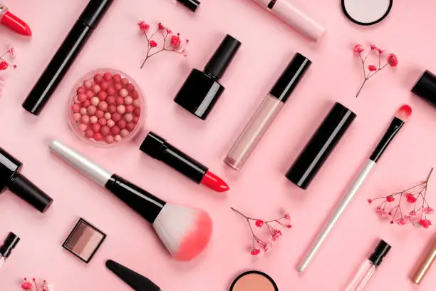 Photo of Various cosmetic accessories for makeup and manicure on trendy pastel pink background with red flowers. Blush, brush, eye shadow, mascara, perfume, lipstick, nail Polish. Skin care products.