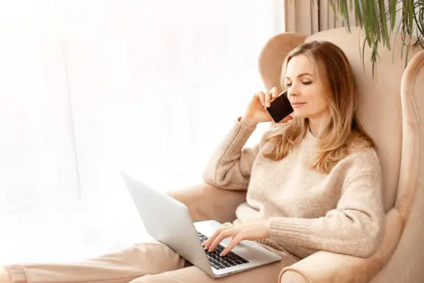 Beautiful young woman typing on laptop and call phone sitting in a chair near the window. The concept of working home office,online shopping, freelancer work,web surfing information,technology concept