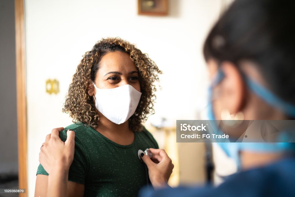 Doctor listening to patient's heart at home during home visit - wearing protective face mask Protective Face Mask Stock Photo