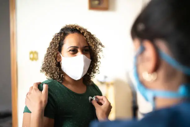 Photo of Doctor listening to patient's heart at home during home visit - wearing protective face mask