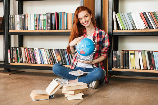 Smiling teenage girl holding globe while sitting at the library