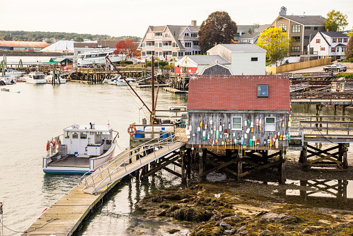 Fishing boats moored to jetties in a harbour along a river at sunset in autumn. A wooden fishing hut with buoys hanging on the exterior wall is in foreground. Kittery, ME, USA.