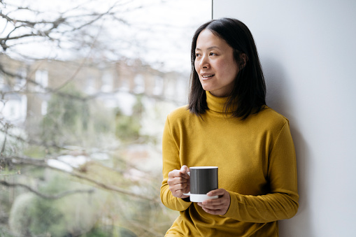 Waist-up view of mature woman with medium-length black hair in gold colored turtleneck sitting on sill and smiling as she holds mug and looks out window.