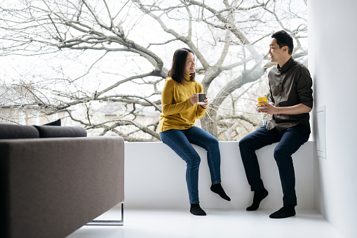 Full length view of man and woman in casual clothing sitting face to face on window sill of modern home enjoying conversation and hot beverages.