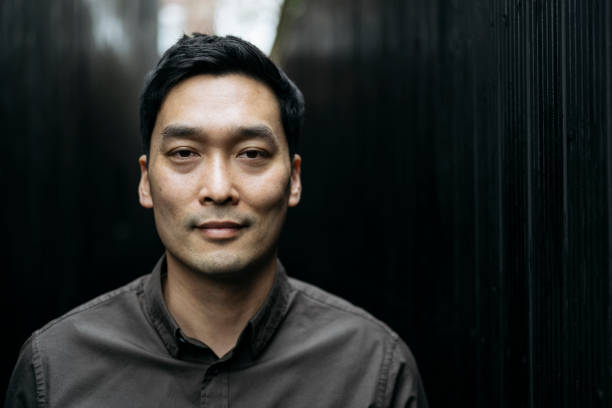 Head and shoulders portrait of confident Chinese man Front view of early 40s man with short black hair in brown button down shirt standing outdoors in natural light and looking at camera with assured expression. low lighting stock pictures, royalty-free photos & images