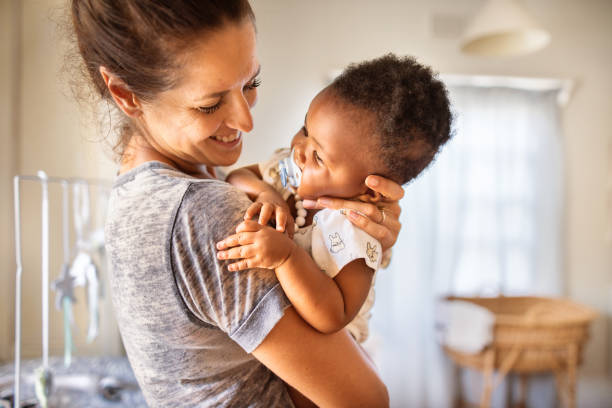 Baby girl with a pacifier being held by her loving mom at home Smiling mom holding her adorable little adopted baby girl with a pacifier in her mouth in her arms at home adoption stock pictures, royalty-free photos & images