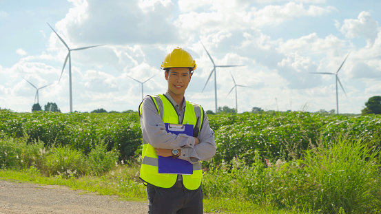 Portrait of Asian windmill engineer man, worker smiling, working on site at wind turbines field or farm, renewable clean energy source. Eco technology for electric power. industry environment. People