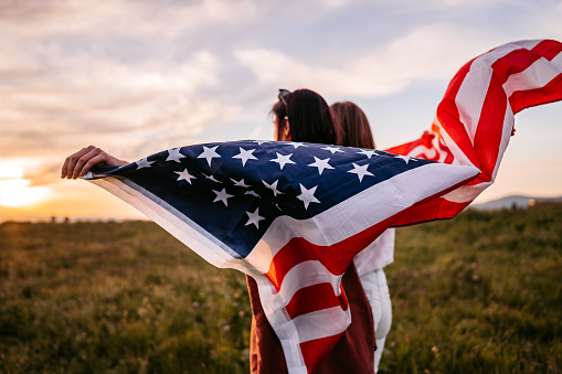 Two, young, females, covering themselves with USA flag. Standing on the meadow at sunset. Rear view.