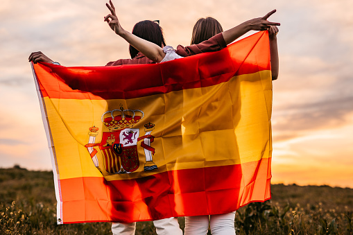 Two, young, females, covering themselves with Spanish flag. Standing on the meadow at sunset. Rear view.