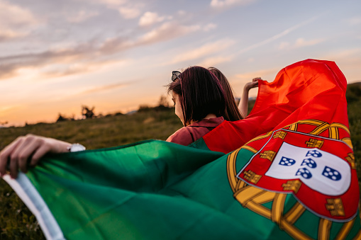 Two, young, females, covering themselves with Portuguese flag. Standing on the meadow at sunset. Rear view.
