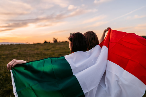 Two, young, females, covering themselves with Italian flag. Standing on the meadow. Rear view.