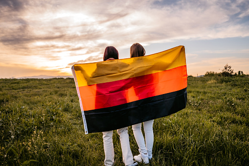 Two, young, females, covering themselves with German flag. Standing on the meadow at sunset. Rear view.