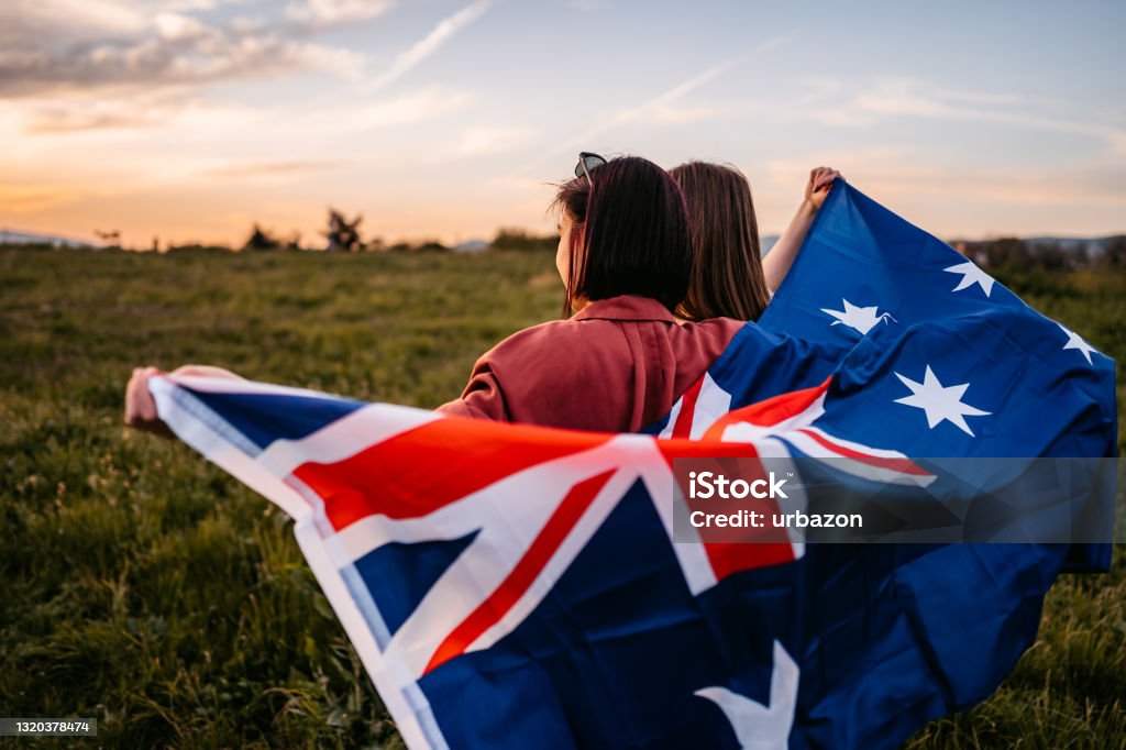 Two women  covering themselves with flag of New Zealand on meadow Two, young, females, covering themselves with flag of New Zealand. Standing on the meadow at sunset. Rear view. New Zealand Flag Stock Photo