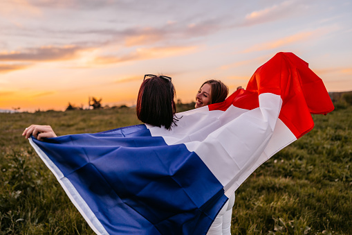 Two, young, females, covering themselves with flag of France. Standing on the meadow at sunset. Rear view.