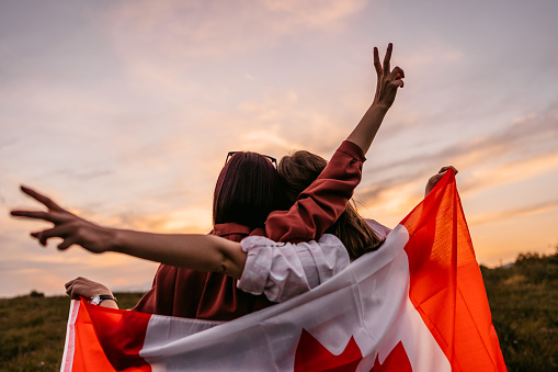 Two, young, females, covering themselves with Canadian flag. Standing on the meadow at sunset. Rear view.