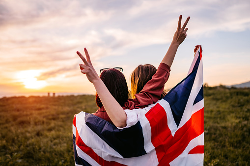 Two, young, females, covering themselves with British flag. Standing on the meadow at sunset. Rear view.