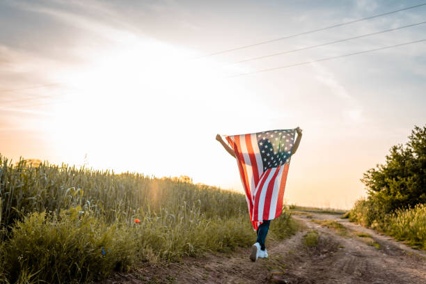 silhouette de femme tient un drapeau américain dans ses mains, levé au-dessus de sa tête. jour de l’indépendance. vue arrière. en arrière-plan, coucher de soleil. - jour de lindépendance photos et images de collection