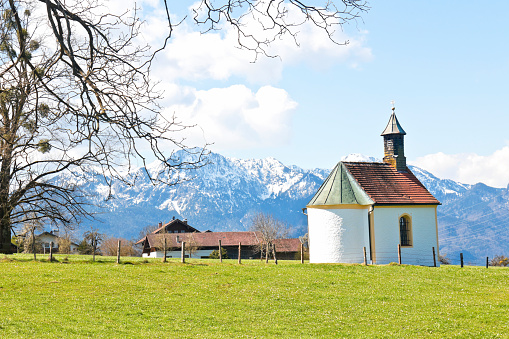 Small chapel with alpine panorama in spring, snowcapped mountains in the background.