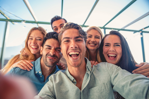 Group of friends taking a selfie laughing and smiling and pulling silly faces. They are standing outdoors on  a balcony