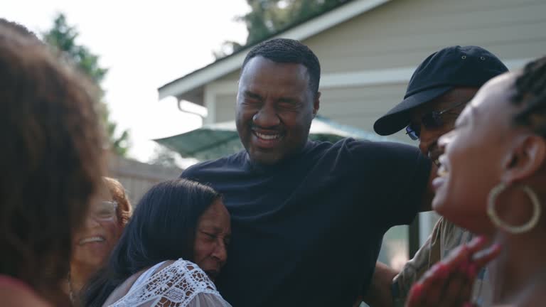 A large multi-generational group of family relatives greets and embraces in the backyard having not seen each other for a long time, due to normal circumstances or perhaps COVID-19 or Coronavirus.  Happiness and joy abound as they are together again.