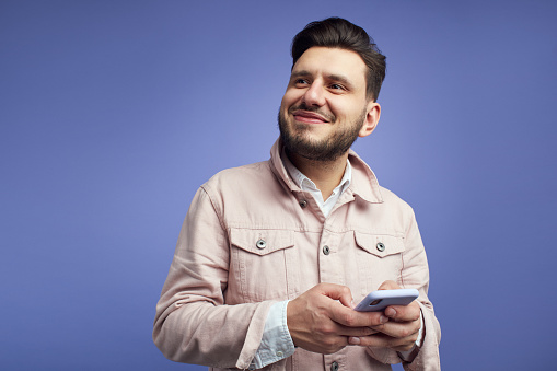 Smiling cheerful hipster with trendy haircut, holds mobile phone, uses app for online communication, concentrated away, isolated on green background