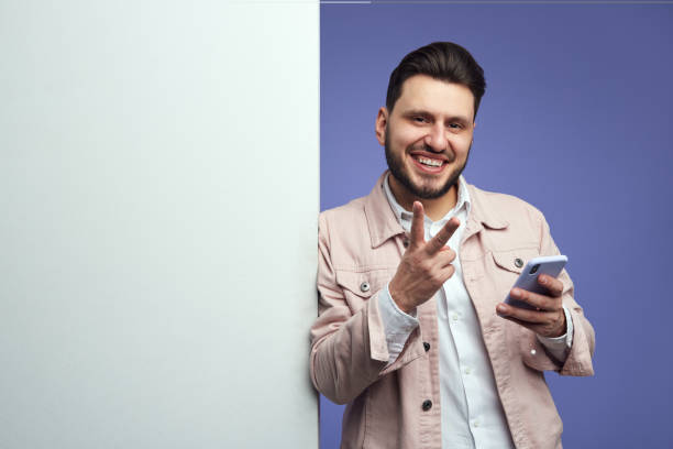 istock Man standing alongside empty white billboard wall and showing peace gesture 1320389598