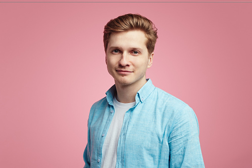 Delightful man in blue shirt, look pleased at camera, has good mood. Fashionable pleasant looking male student poses over pink background
