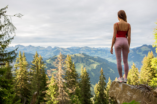 Relaxed young woman hiker standing high above on top a mountain looking at the beautiful green nature forest landscape view.